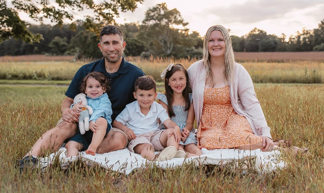 Two parents and three children sitting on a blanket in a grassy field with trees in the background.